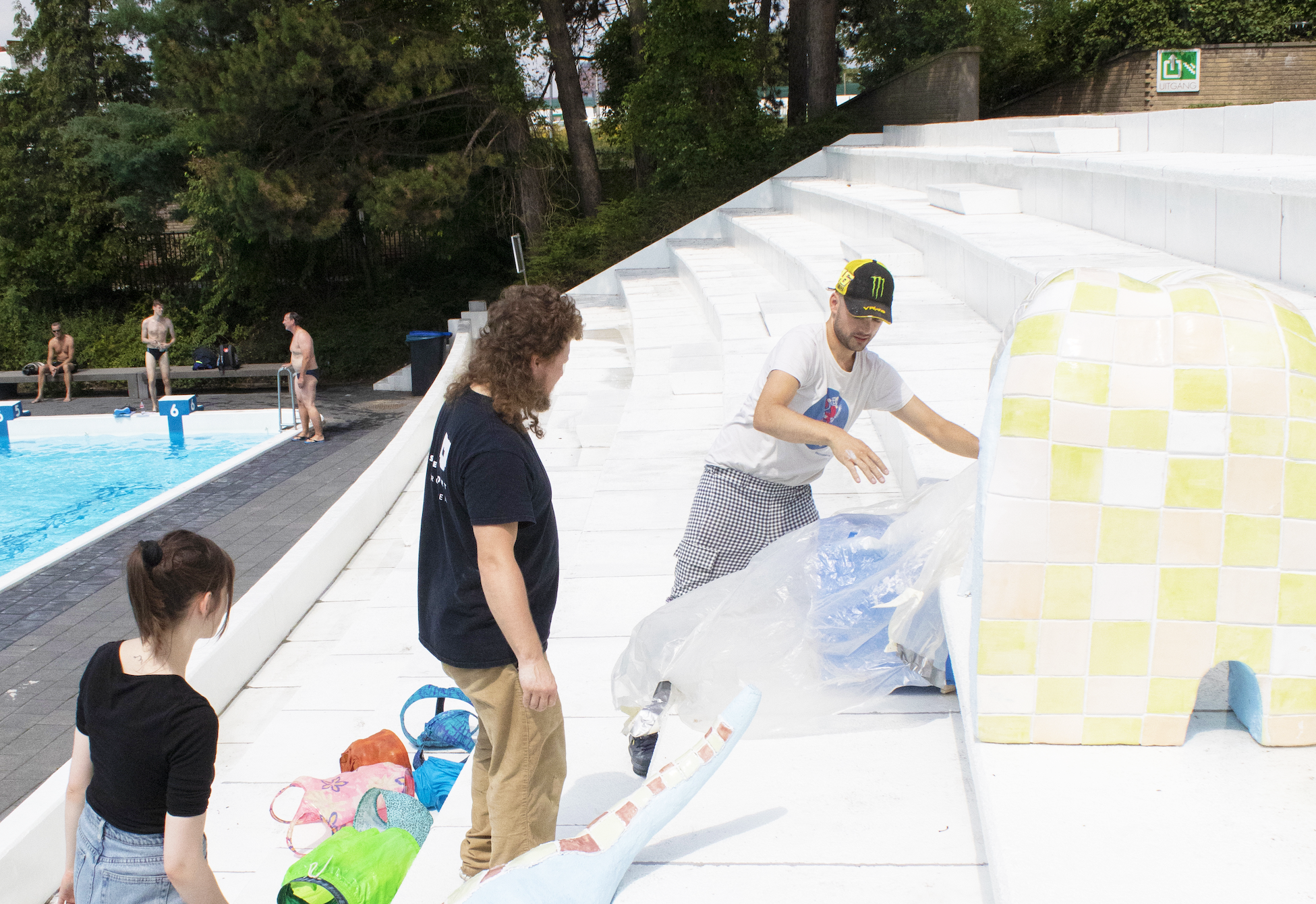 Drie personen staan op de trap naast een zwembad. Een van de personen legt een stuk plastic op een kunstwerk.