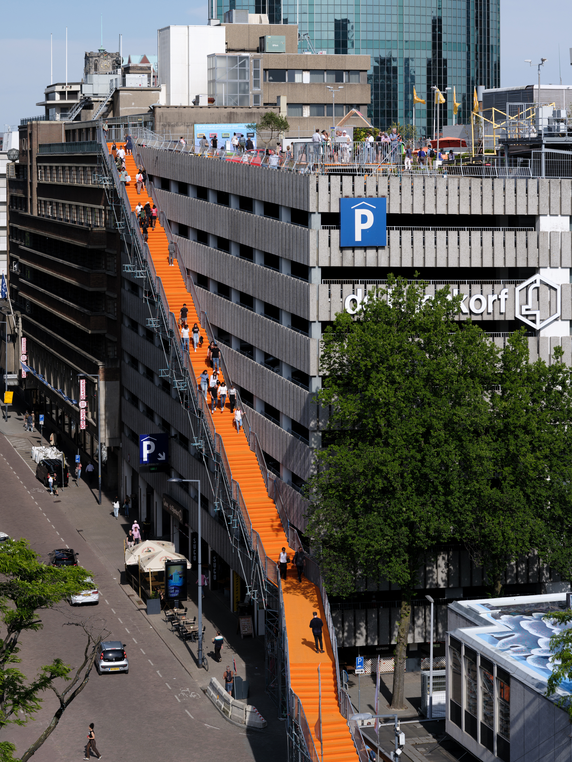 personen lopen via een oranje trap het dak op van de Rotterdamse Bijenkorf