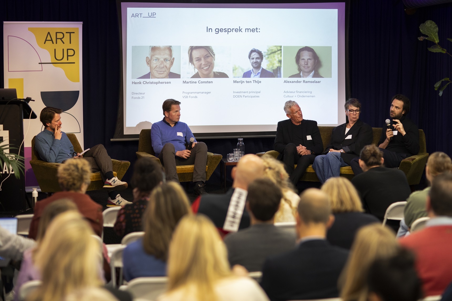 Voorgrond: achterhoofden van publiek. Achtergrond: moderator en vier panelleden op een podium.