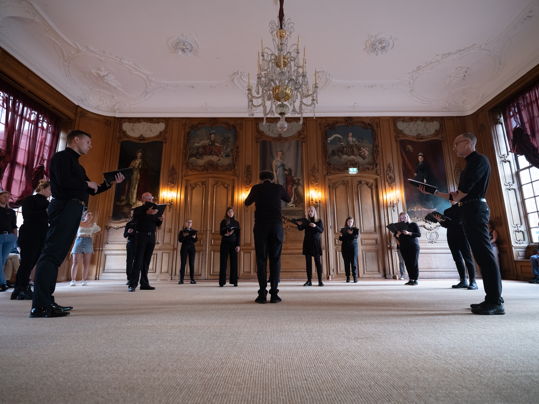 kamerkoor bestaande uit 12 mensen in zwarte pakken zingen in een formele zaal