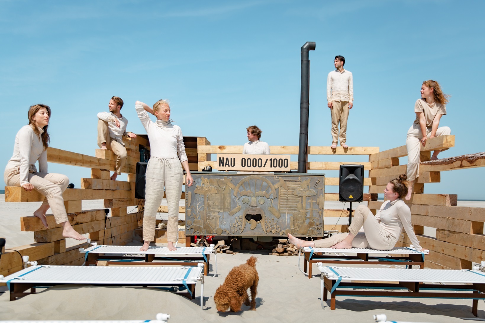 theatergroep van zeven mensen in zandkleurige kleding, een bruine hond, op het strand met een strakblauwe hemel