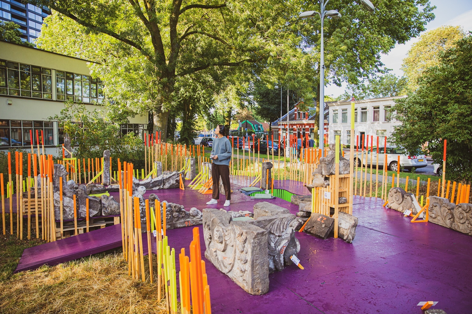 Een vrouwelijk bezoeker beluistert een audiotoer, op een paarse plattegrond met oranje piketpaaltjes en betonnen fundamentstenen.