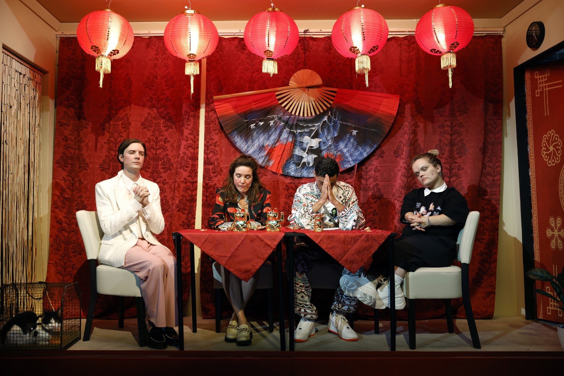 Vier mensen zitten aan tafel met hun handen in gebedshouding. Aan het plafond hangen lampions en op de achtergrond hangt een waaier.