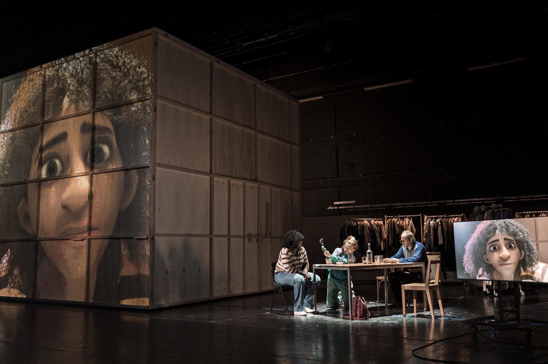 Drie personen zitten aan tafel op een podium naast een houten huis waar een projectie van een vrouw op te zien is