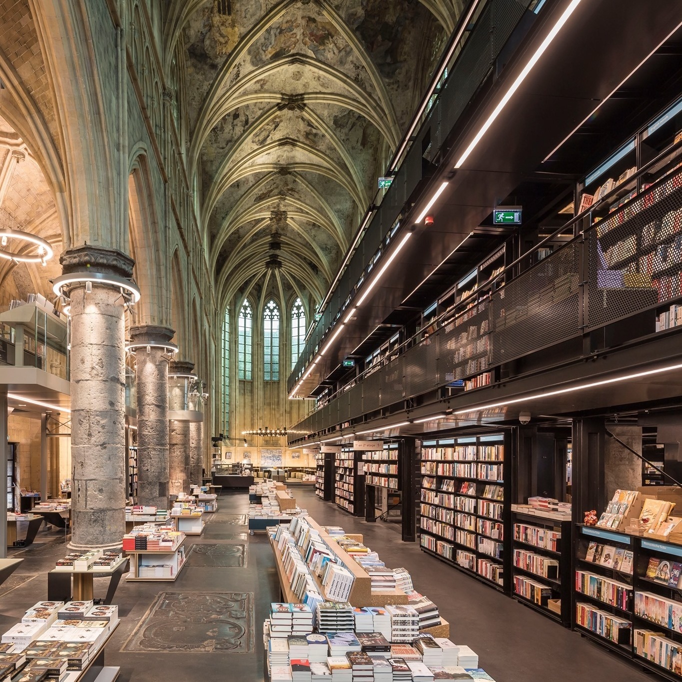 Interieur Dominicanen Boekhandel. Voormalige kerk in Gotische stijl met hoge pilaren en versierde, boogvormige plafonds. De boeken liggen op tafels opgesteld op de begane grond en rechts in een hoge kast met verdieping.