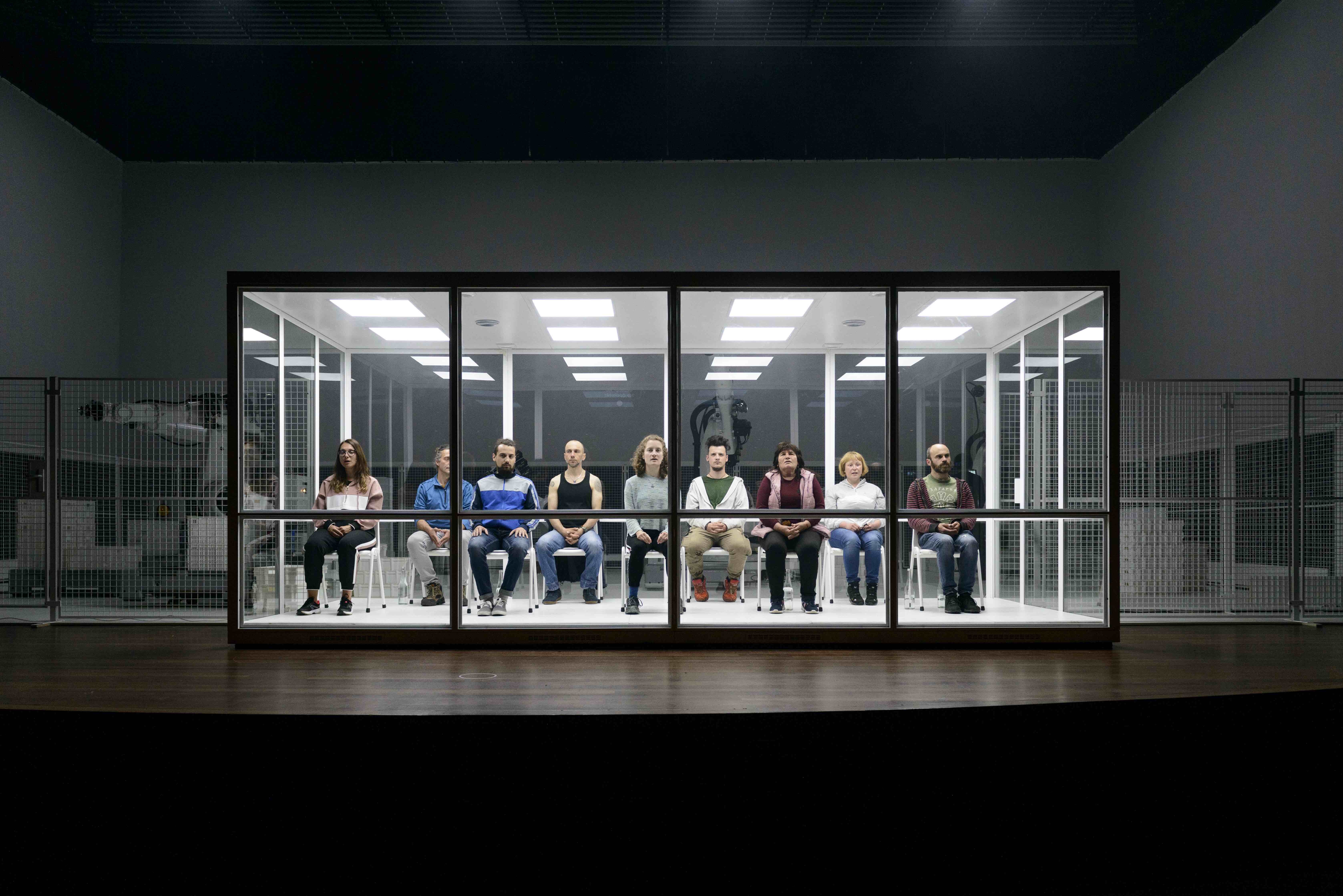 Negen verschillende personen/acteurs, qua gender en leeftijd, zitten in een TL-verlichte glazen ruimte op een rijtje. allen zitten op een wit plastic stoeltje.