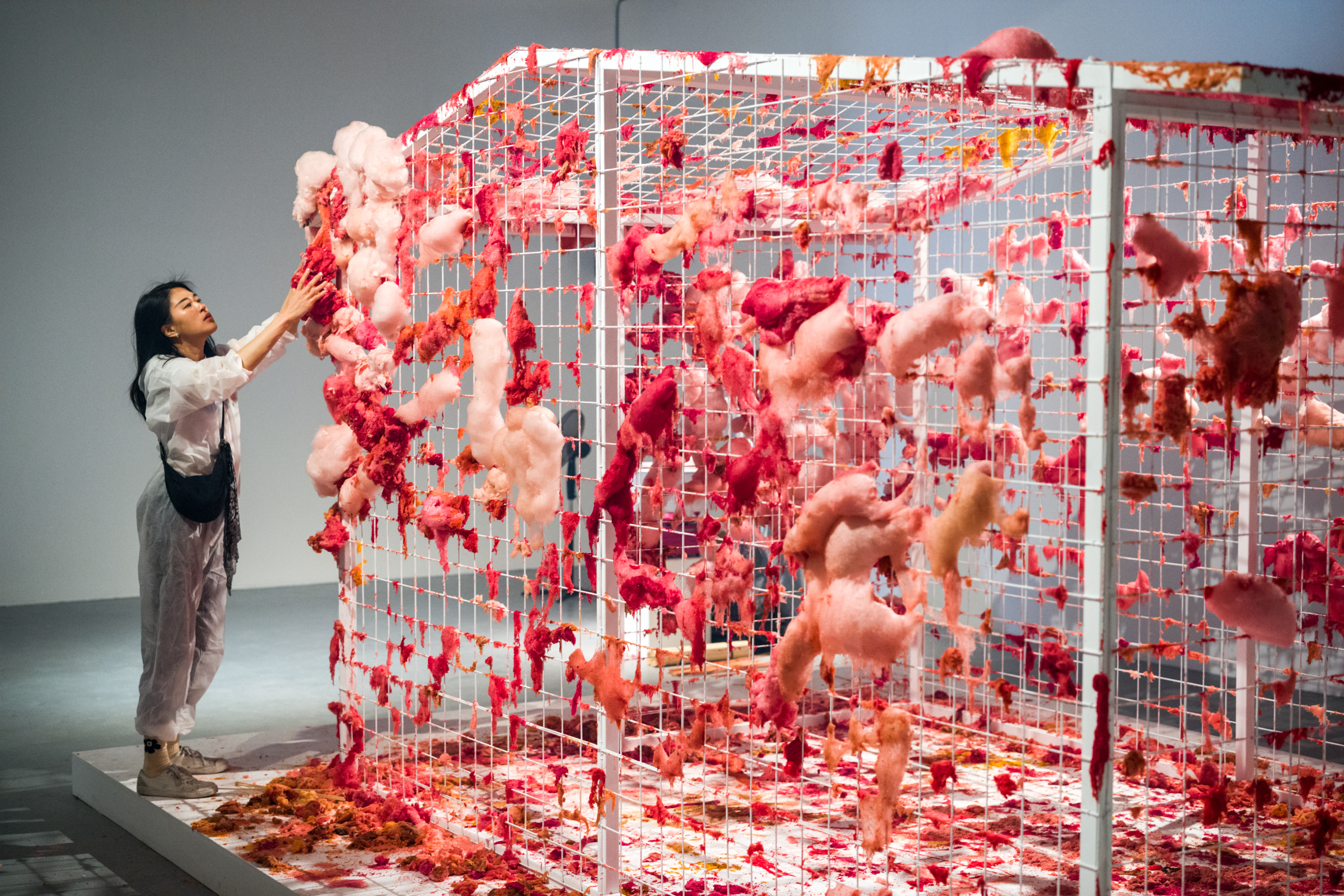 Een metalen witte kooi met vierkante raster hangen roze kleurige schuimen elementen. Een vrouw in een wit pak houdt een van deze vormen vast.