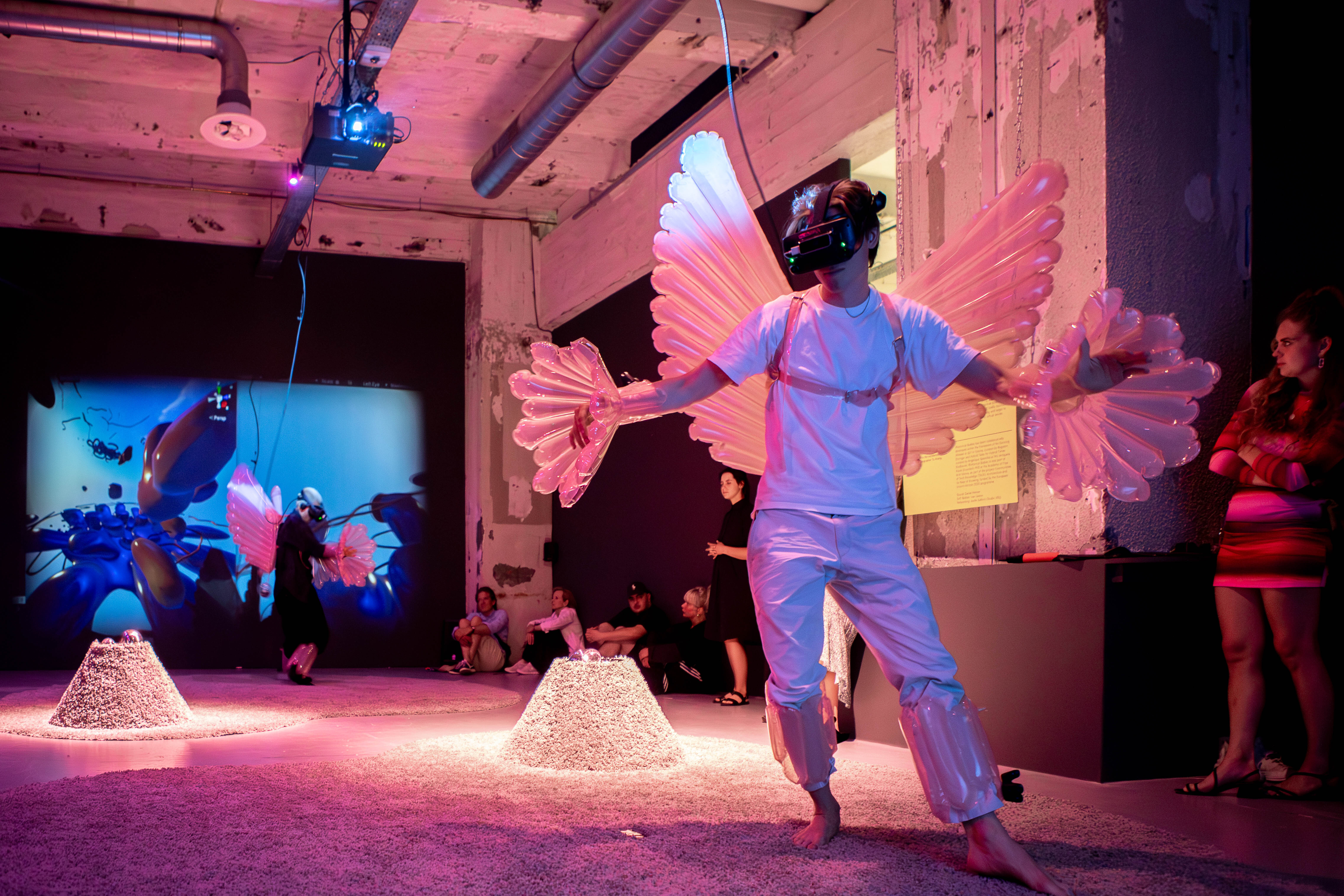 In een tentoonstellingsruimte staat een persoon met een vr-bril op en roze opblaasvleugels op diens rug en aan de handen. Achterin is een project te zien en zitten mensen op de grond.