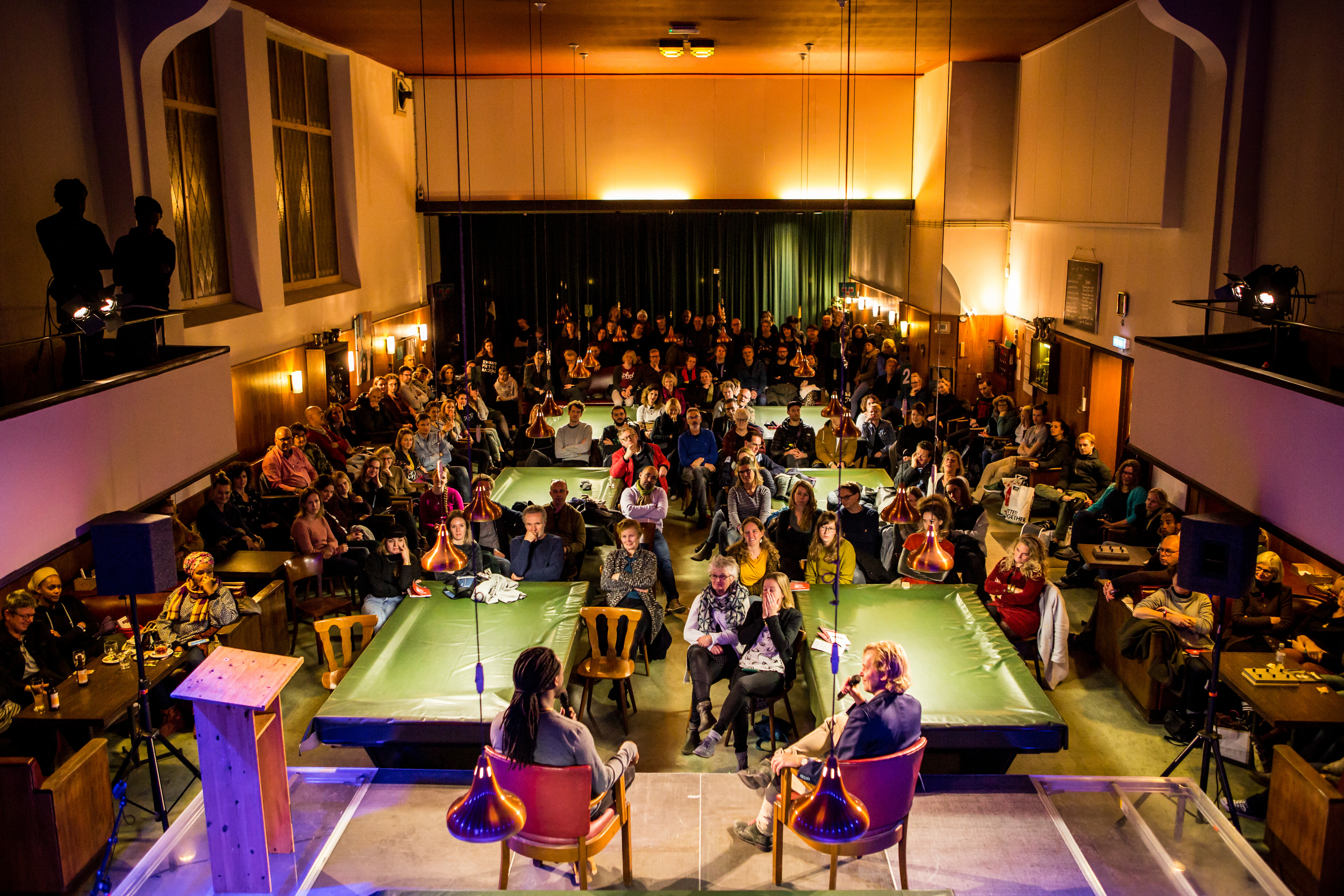 Twee sprekers op een verhoging in een biljartzaal vol met luisteraars