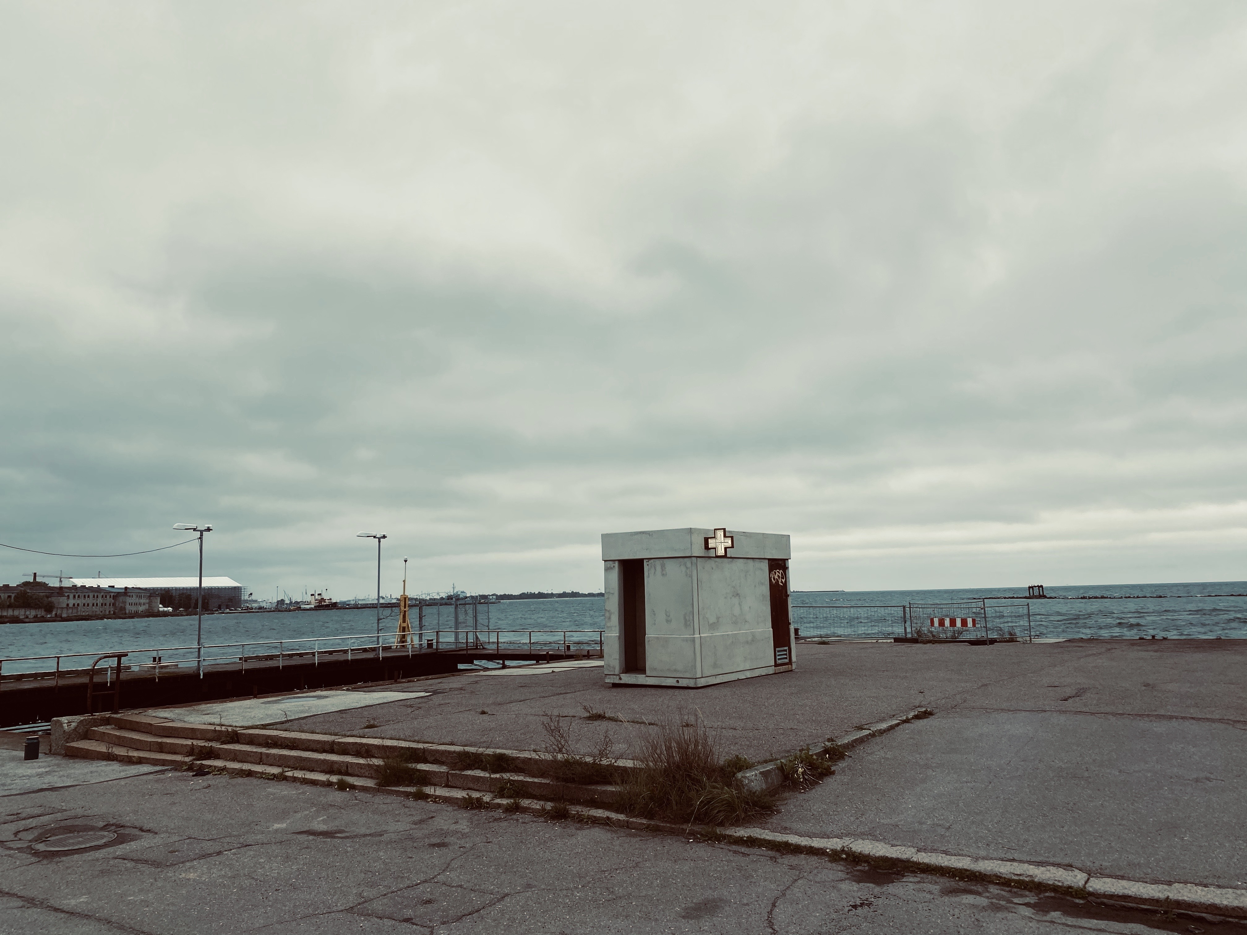 Een klein stenen gebouw, met een verlicht kruis hangend aan de muur. Het gebouw bevindt zich op een verlaten terrein en heeft water op de achtergrond.