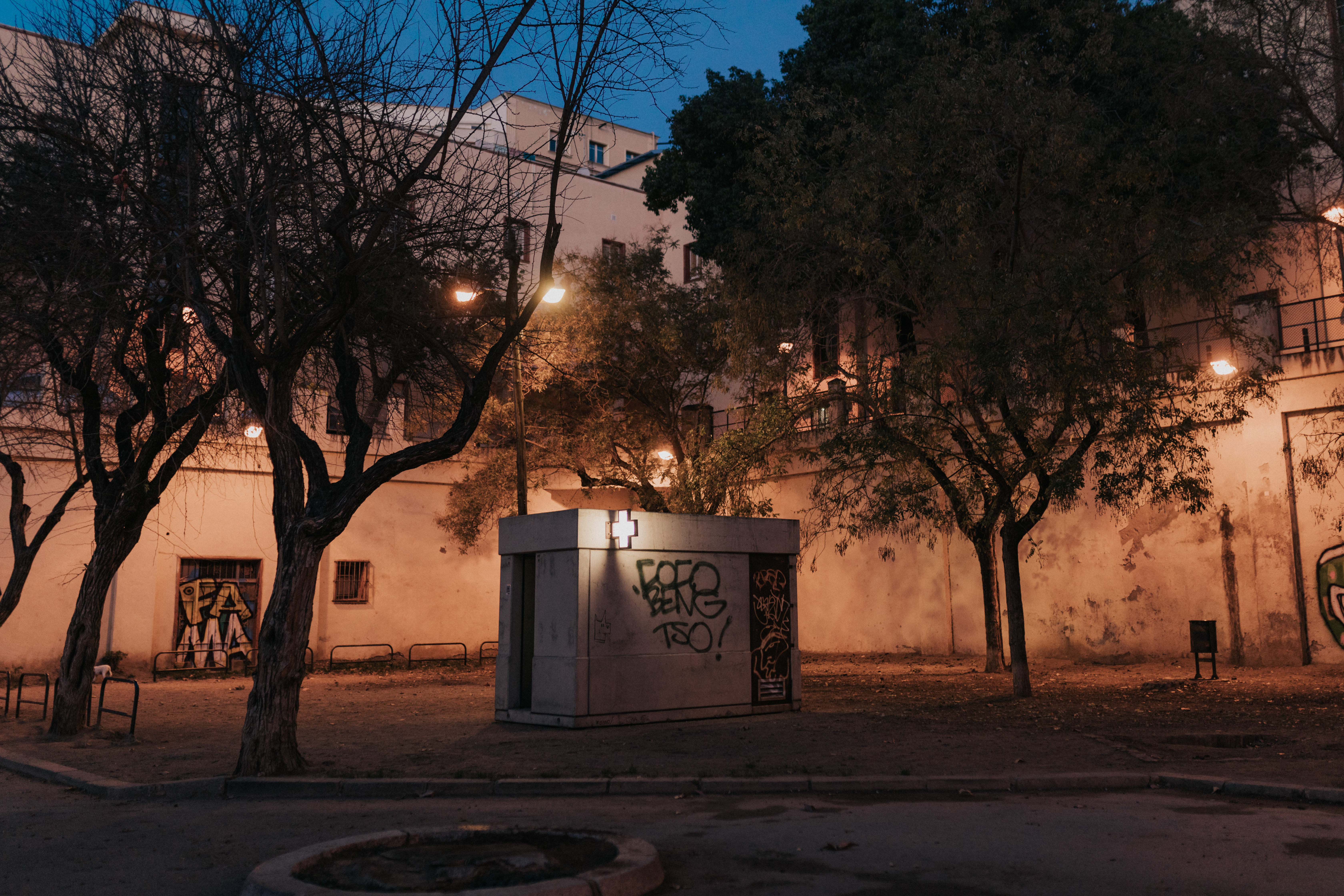 Een stenen gebouw, met graffitti tags en een wit verlicht kruis op de muren, bomen eromheen en een groot wit gebouw achter de bomen. Het is halfduister