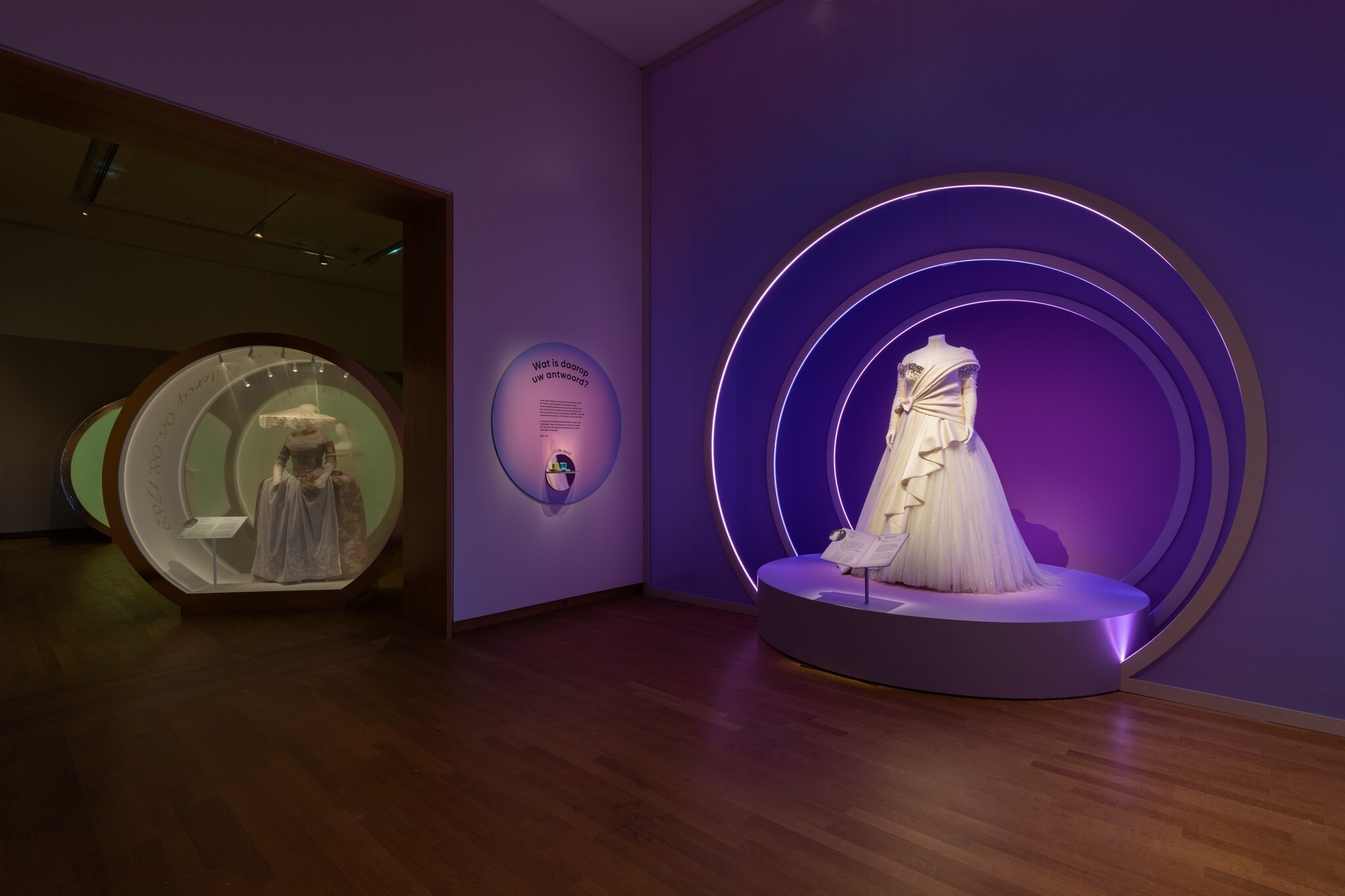 Een tentoonstellingsruimte met twee ruimtes (de eerste paars en de ander groen) waar jurken op een display staan met bogen eromheen. Naast het podium hangt de zaaltekst.