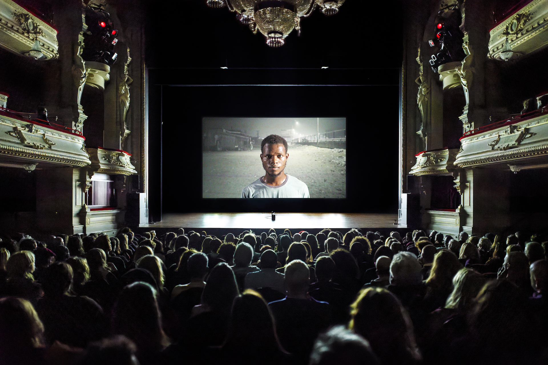 Performer - te zien via een groot podiumvullend scherm - kijkt de klassieke schouwburgzaal van ITA in vanuit Sea Side Cottage in Lagos, Nigeria.