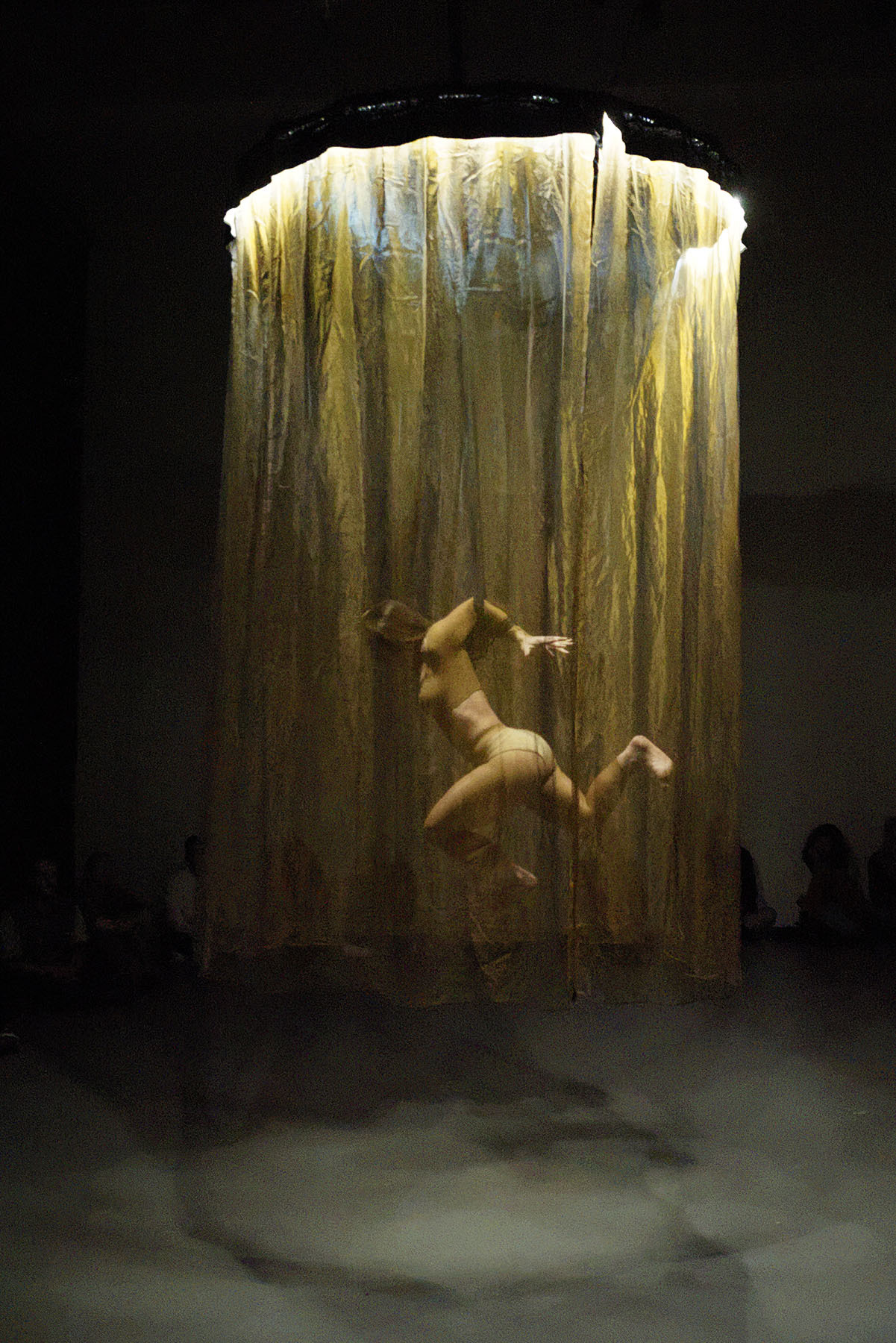 Een vrouwelijke performer/ballerina neemt een danspose aan in een tuigje boven de grond. Er hangt een gele voile om haar heen in een diameter van ongeveer 2 meter.