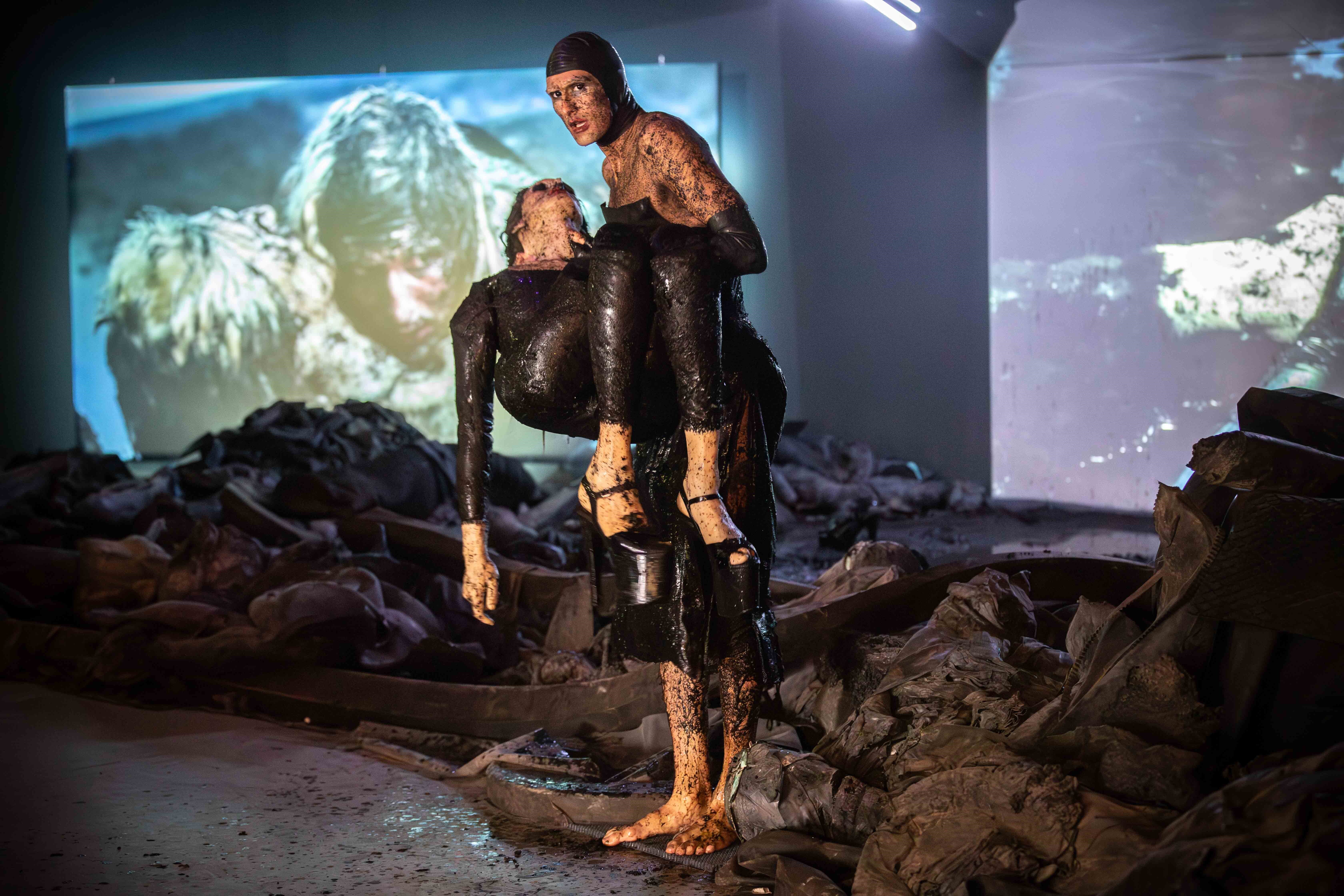 Een man houdt een vrouw vast in zijn armen, beide zitten onder de moder. Op de achtergrond zie je twee beeldschermen. Ze staan in een ruimte waar zwart grijze rotzooi op de grond ligt