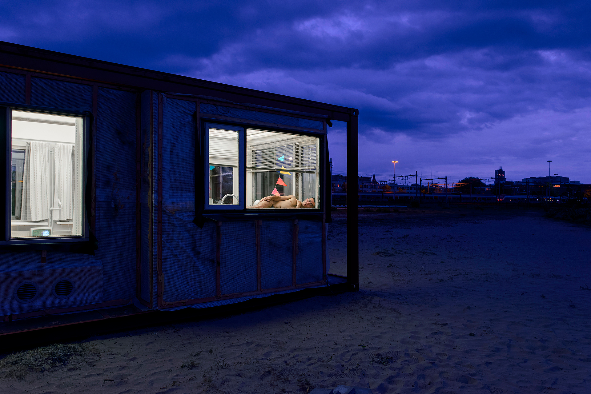 Een container gebouw met achter het verlichte raam een persoon naakt liggend met slingers op de achtergrond, het is bijna donker, de lucht is donkerpaars