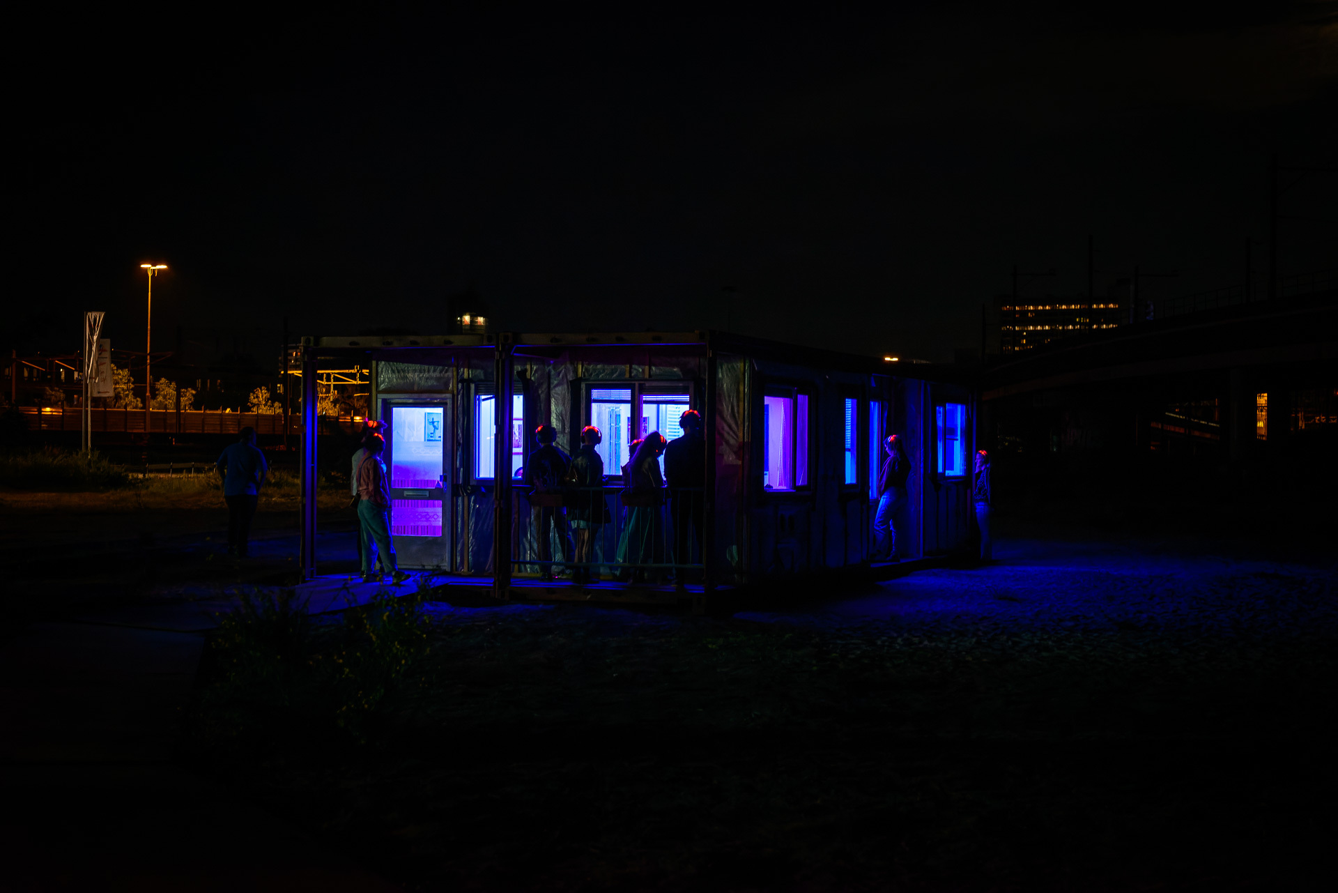 Het is donker en je ziet een gebouw met ramen, die neon paars zijn verlicht. Je ziet de contouren van 8 personen, met rode verlichte hoofden