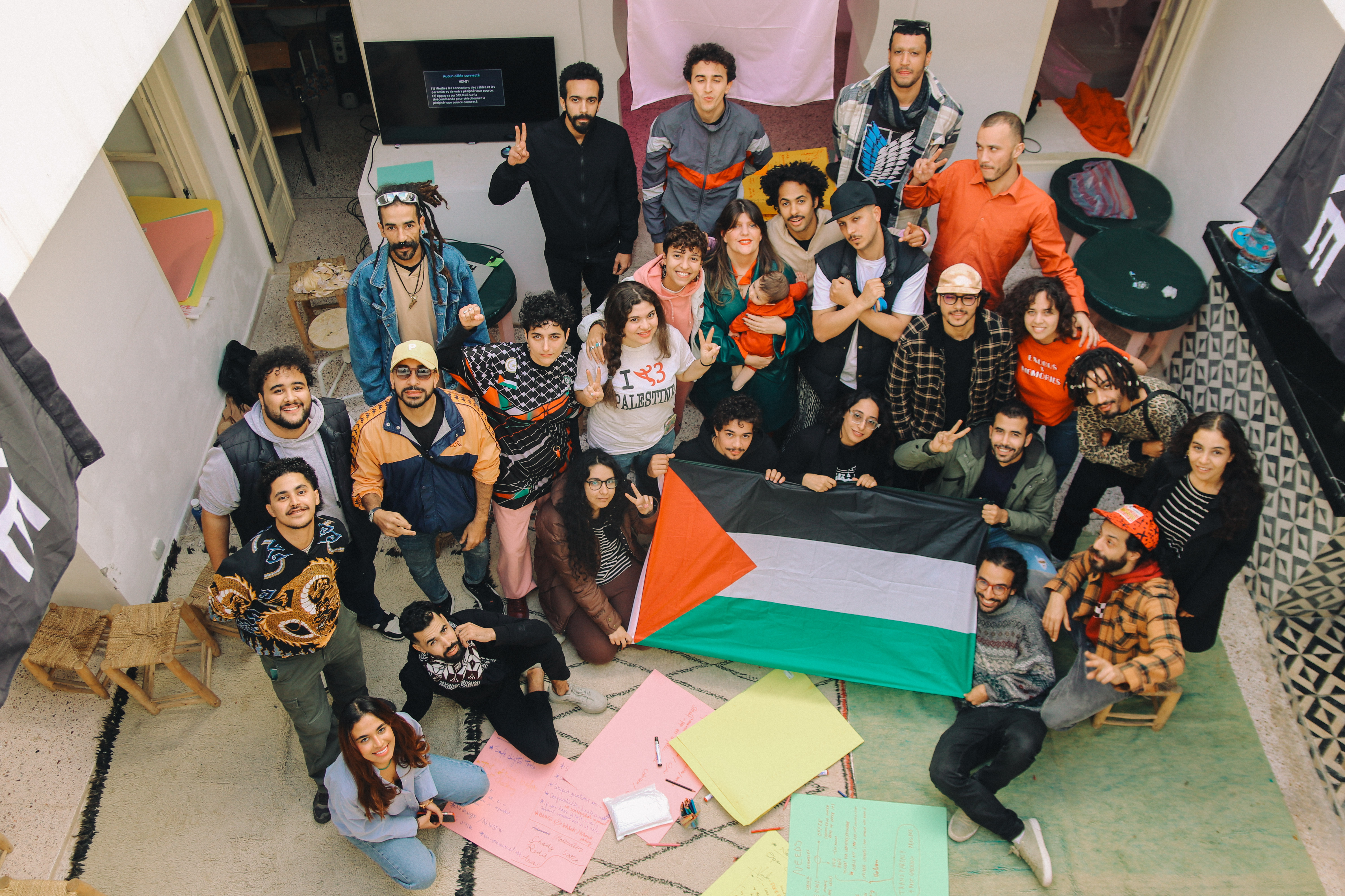 Vogelperspectief op groep van ongeveer 25 mensen die bij een aantal grote vellen papier zitten en staan, en een vlag vasthouden