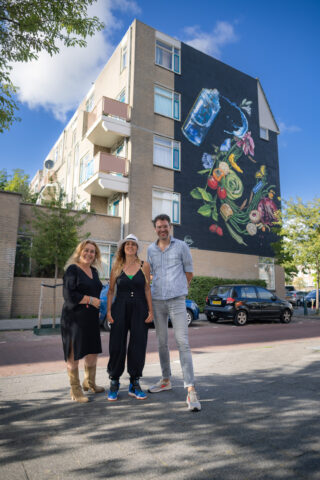 Twee vrouwen en een man staan voor een gebouw met een muurschildering. De muurschildering verbeeldt een graffitti spuitbus waaruit water vloeit over een explosie van ingrediënten en lekkernijen die je kan kopen in het Transvaalkwartier, op de Haagse Markt en in de buurtwinkels.