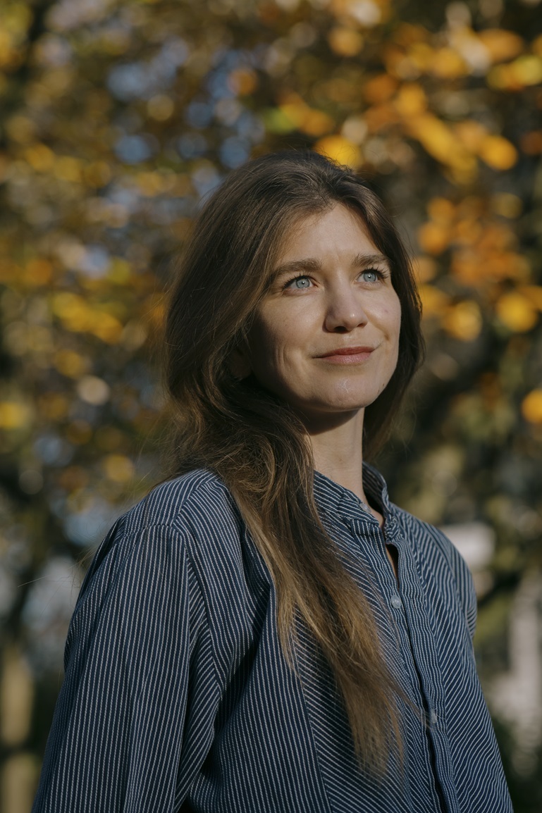portretfoto van vrouw met lang bruin haar. op de achtergrond is vaag een boom in herfstbladeren te ontwaren.
