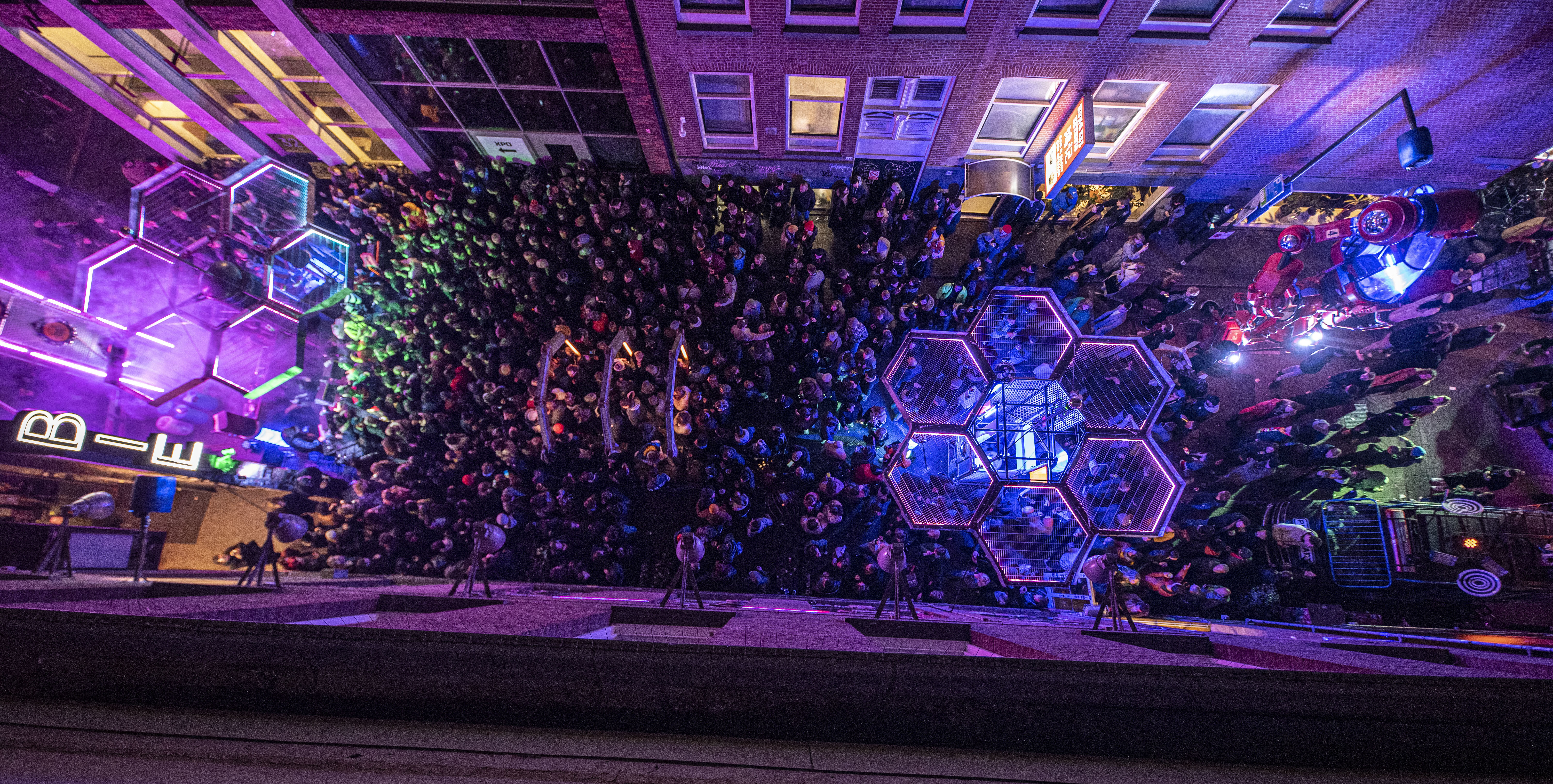 Vogelperspectief op menigte mensen met in het midden een installatie in de vorm van een honingraat. De belichting is paars en groen.